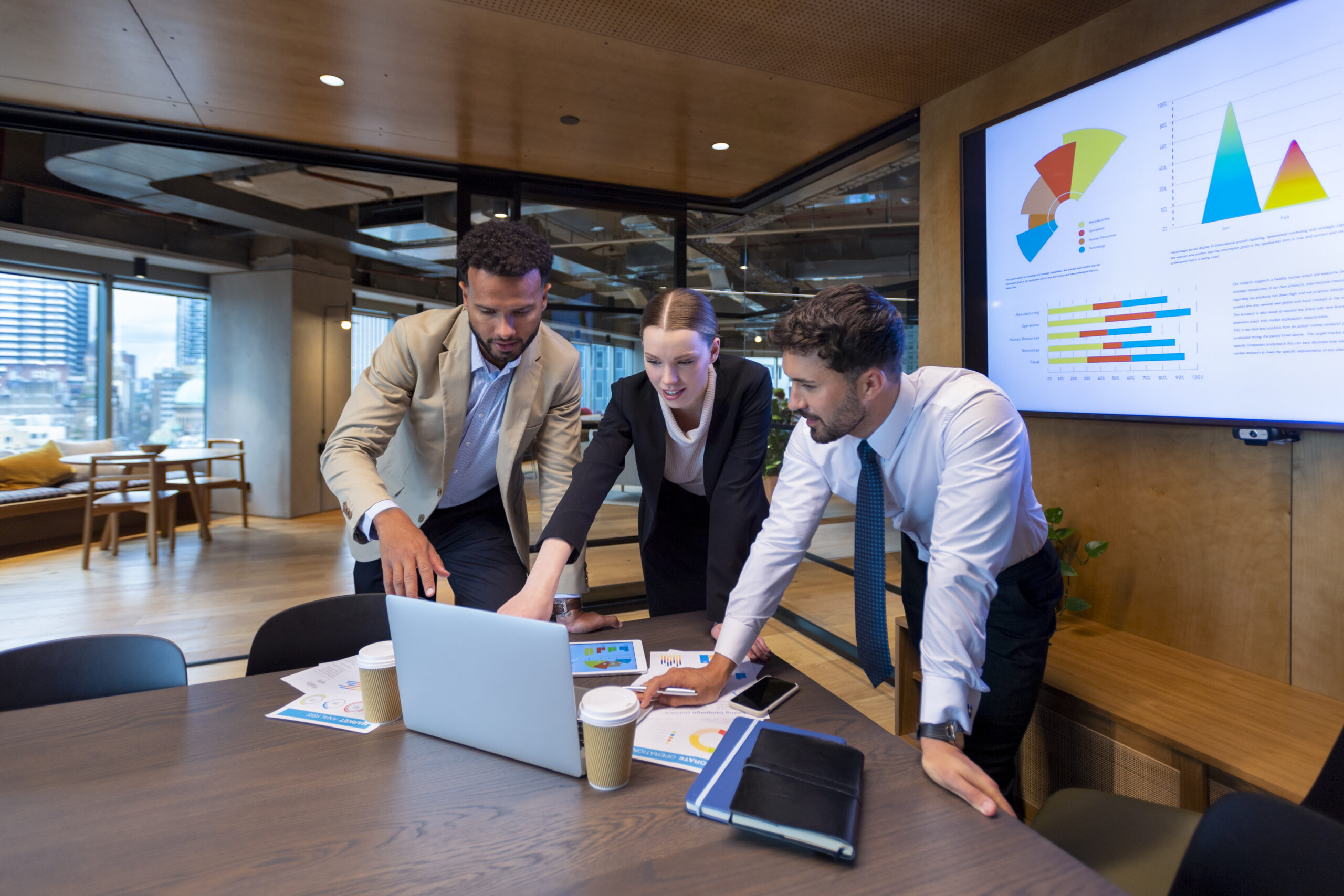 Business people working on a laptop computer in a modern office. There is paperwork on the table with charts and graphs on it. There is a screen behind them with financial charts and graphs on it.