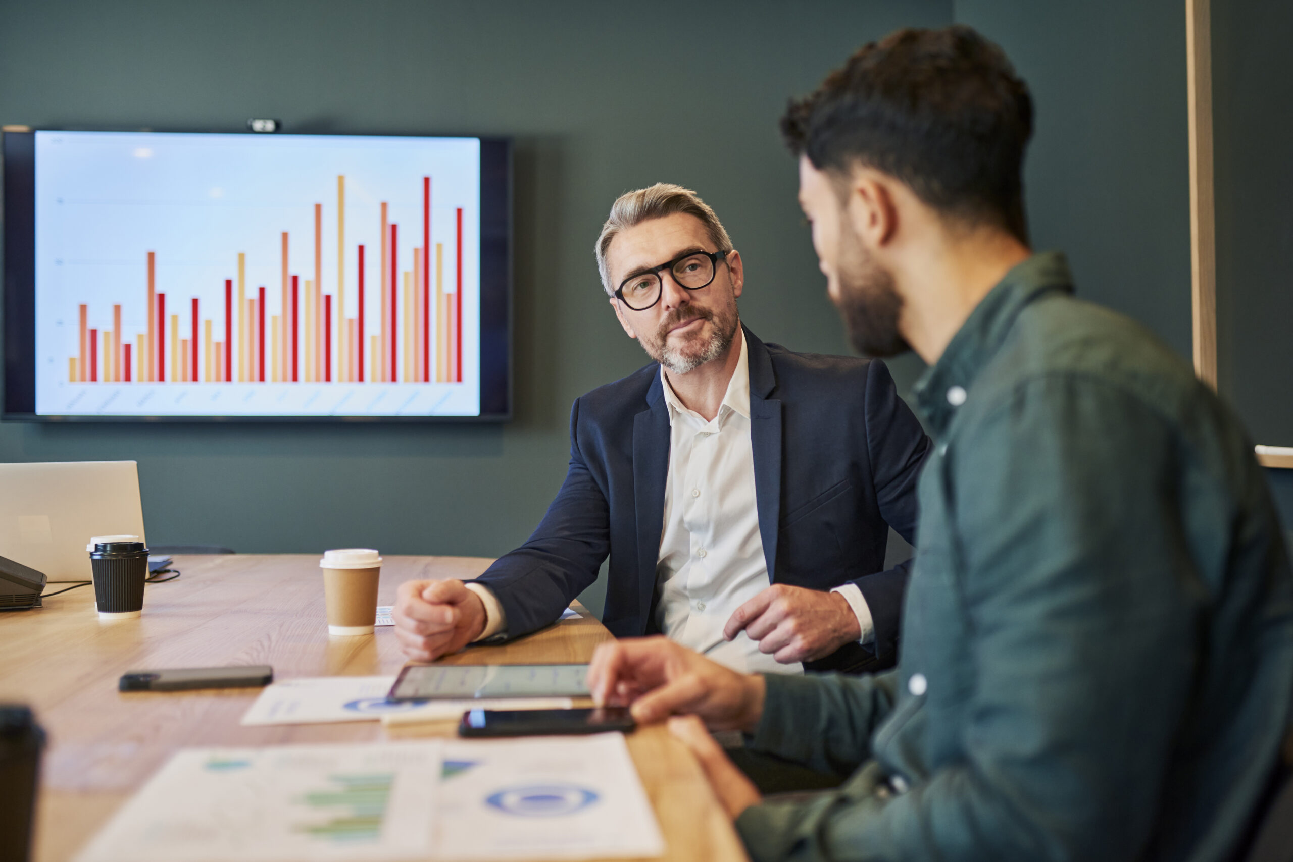 Two business professionals, one older and one younger, are sitting in a conference room having a serious discussion. They are both looking at the younger man's tablet. The older man has a thoughtful expression on his face, while the younger man is looking at him intently.